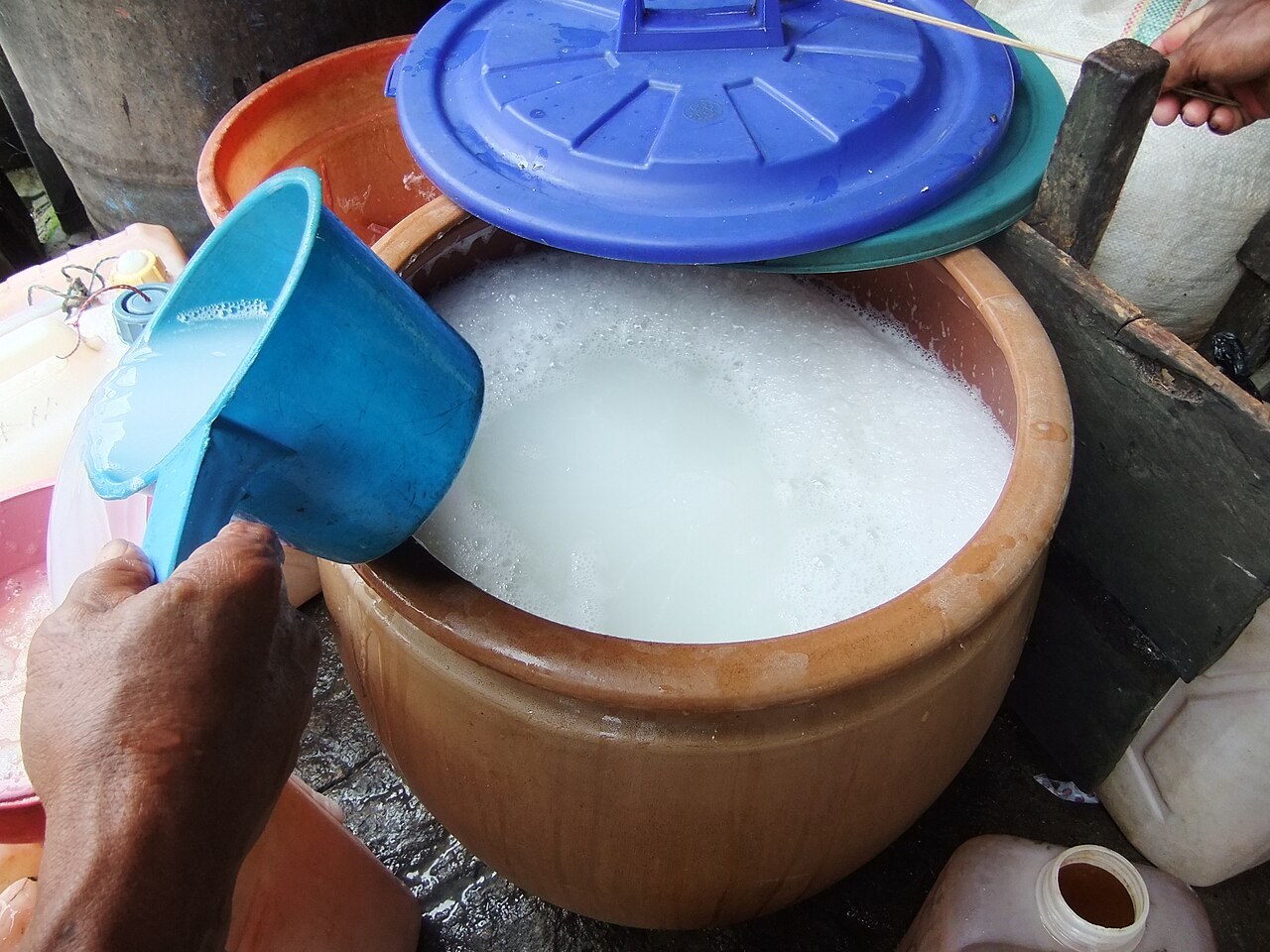 A container of cloudy ballo palm wine being served with a blue scoop